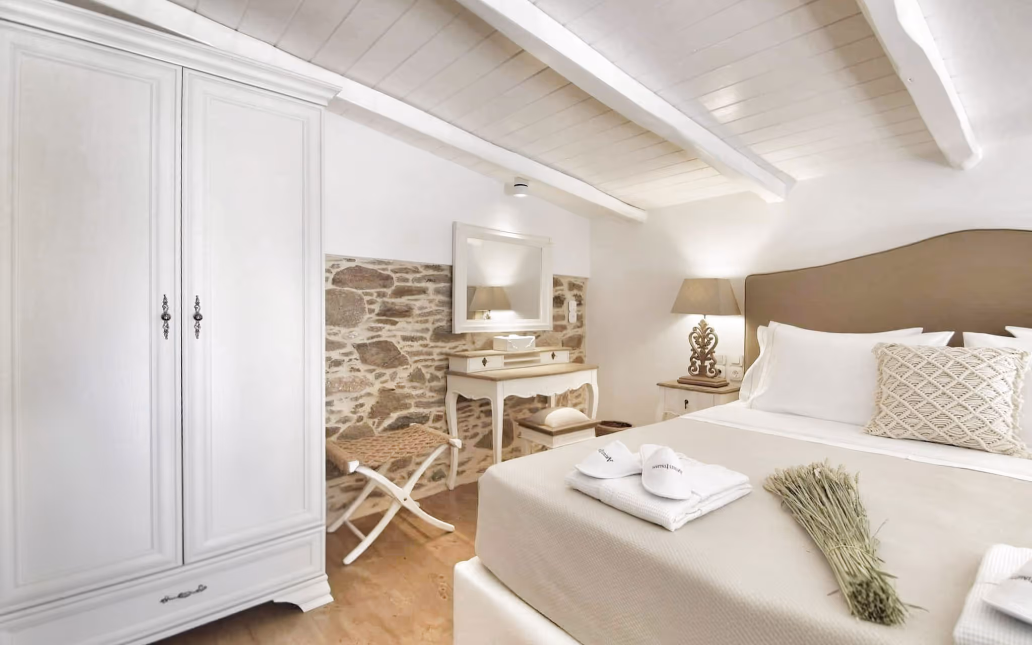 Cozy bedroom with white wardrobe, beige bed, textured pillow, lamp, and rustic stone accent wall.