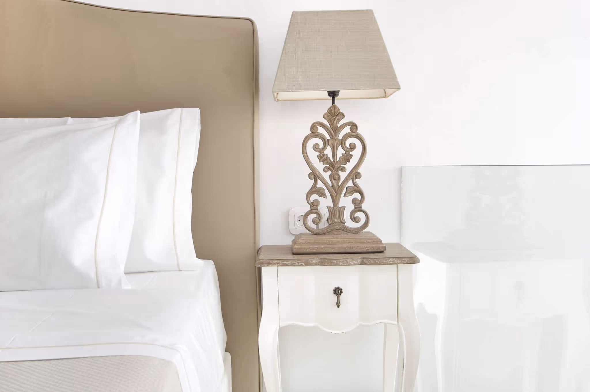 White bedside table with a carved wooden base lamp next to a beige upholstered bed.