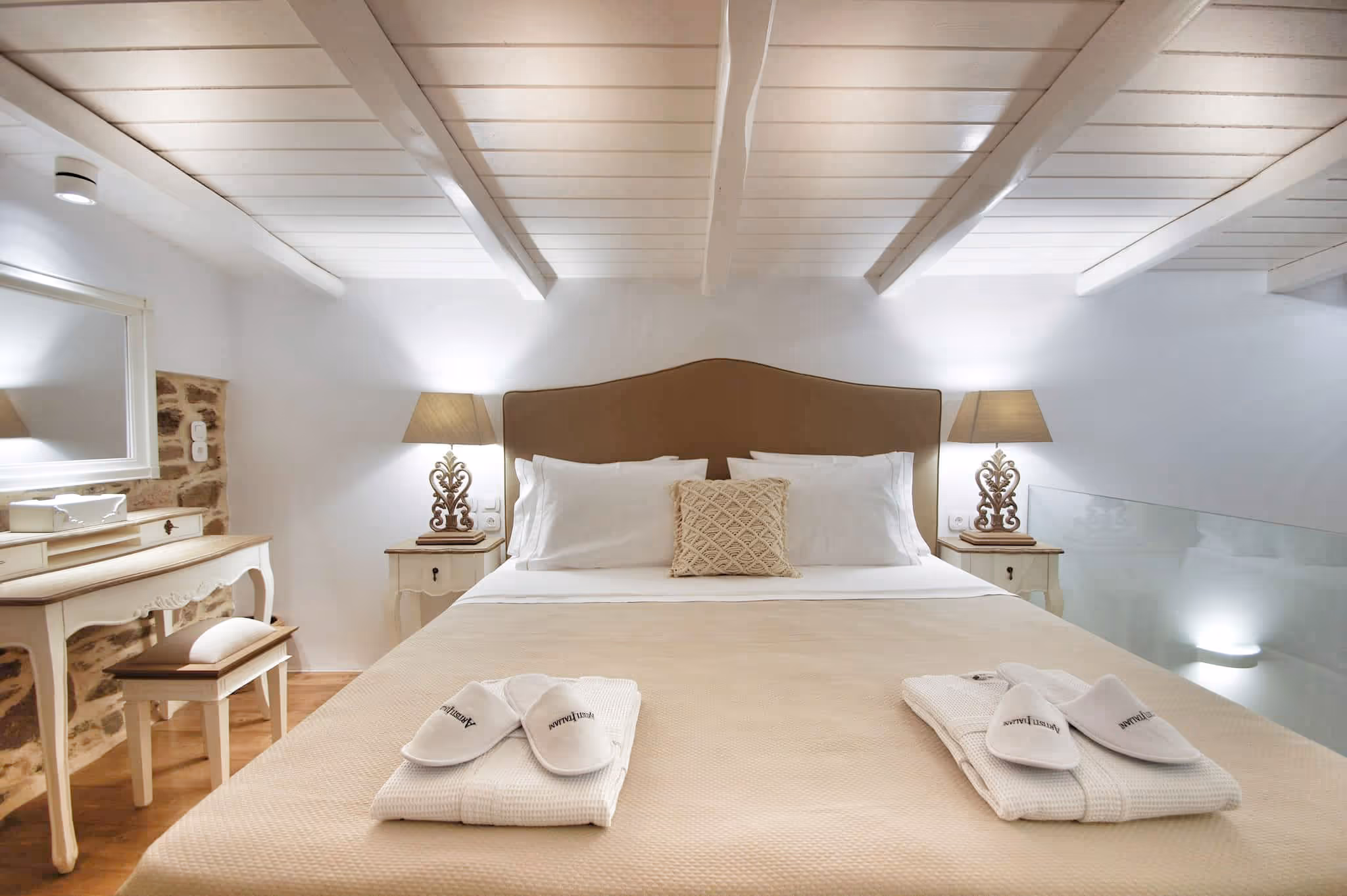 Cozy bedroom with beige bed, white pillows, bedside lamps, and towels with slippers on the bed.
