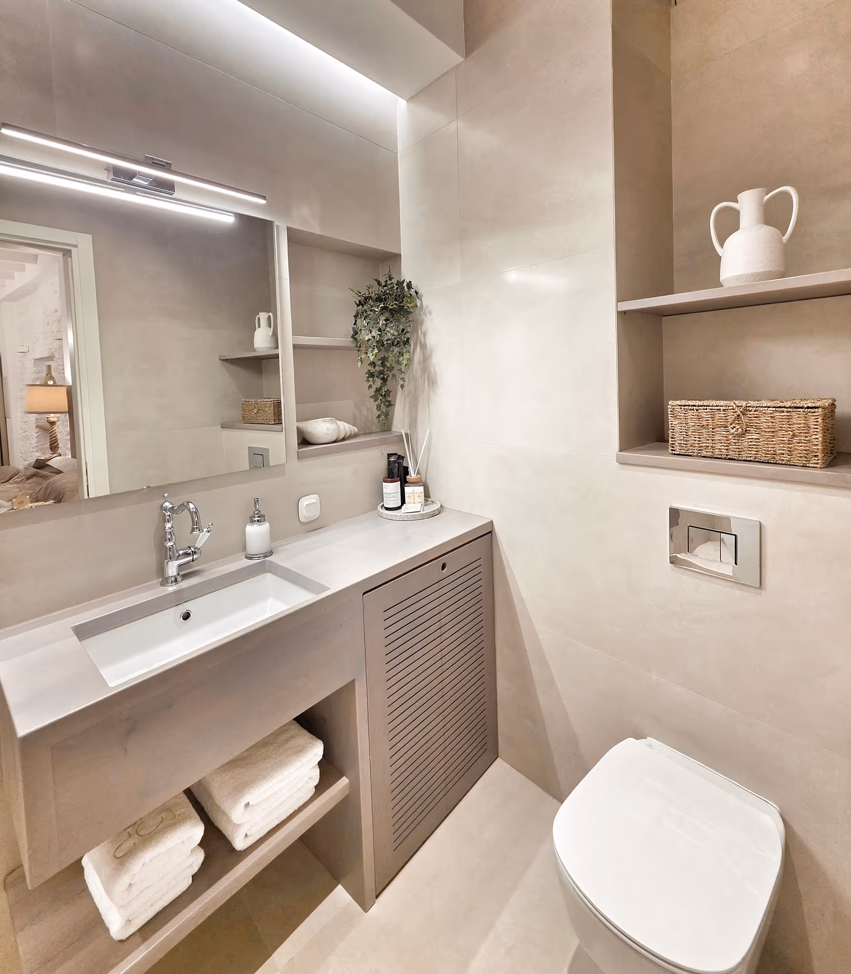 Minimalist beige bathroom with rectangular sink, wall-mounted toilet, shelves with decor and towels.