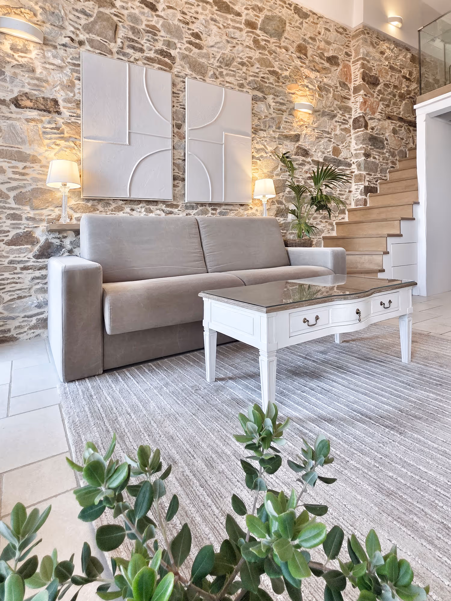 Modern living room with gray couch, white coffee table, stone wall, stairs, lamps, and green plants.