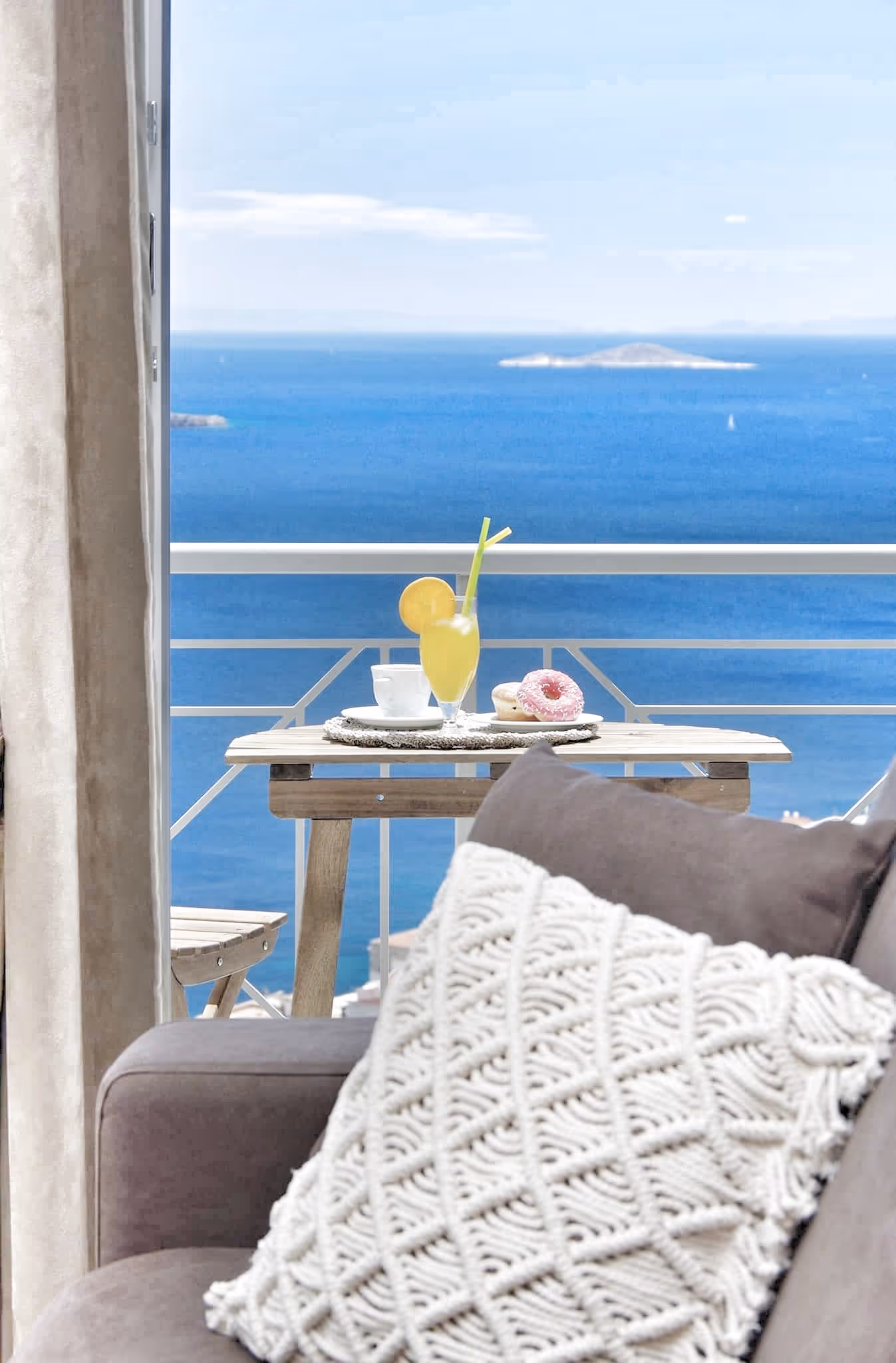 Cozy sofa with white knit pillow facing a balcony table with drinks and donuts overlooking the ocean.