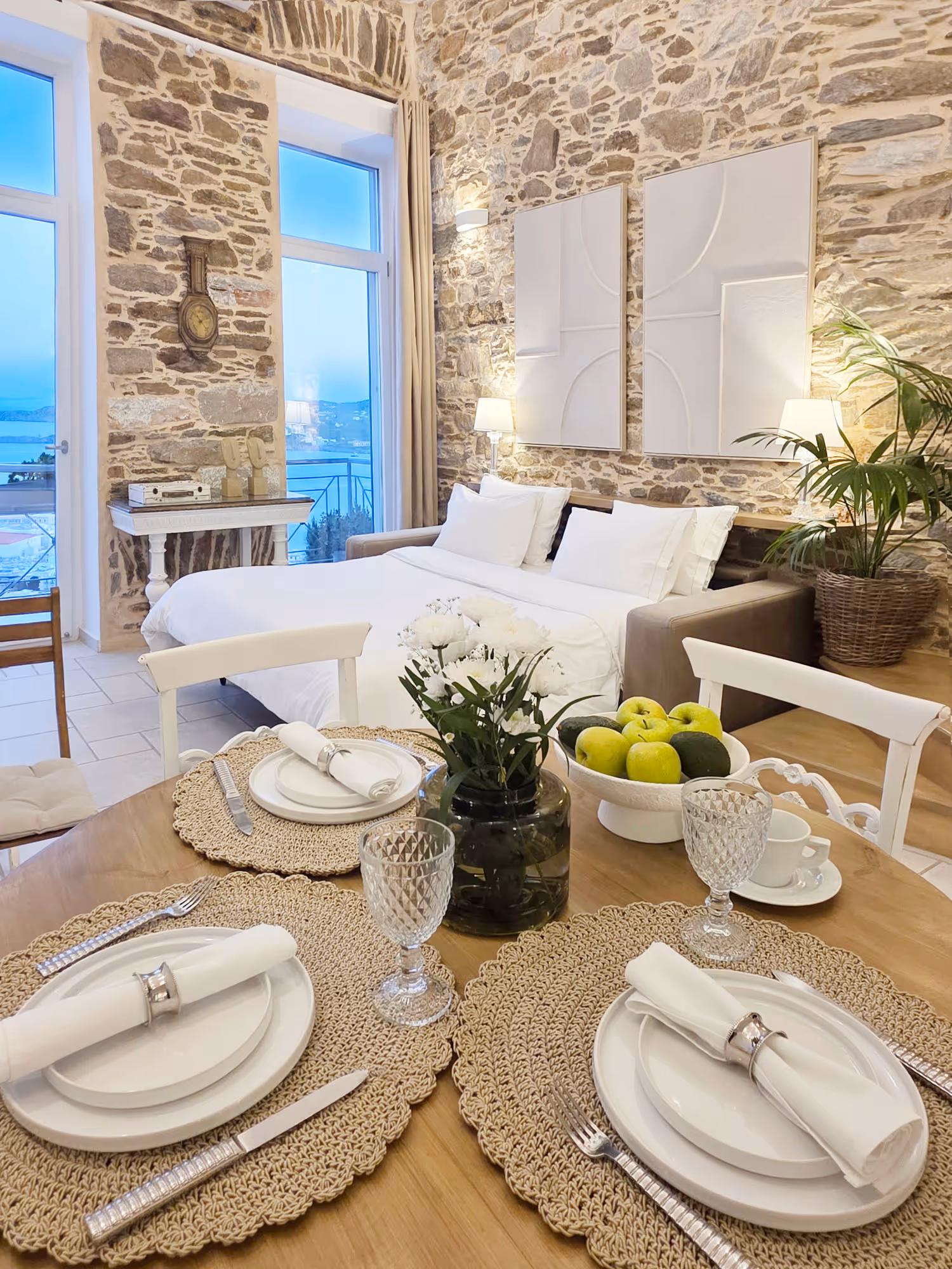 Cozy dining table set with white plates, napkins, glasses, vase with flowers, and green apples in a stone-wall room.