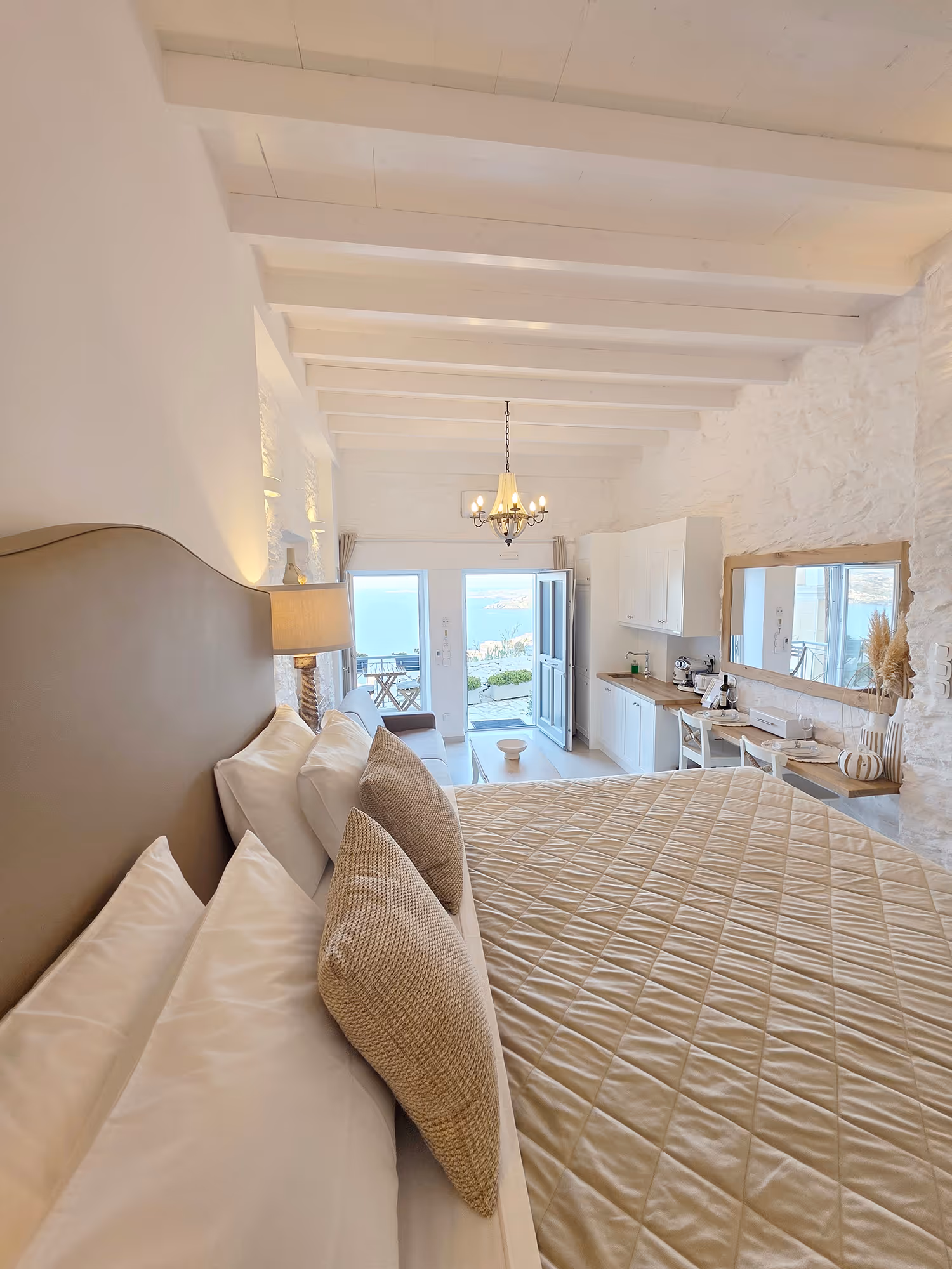 Bright bedroom with beige quilted bed, brown pillows, white walls, desk, and open door to sea view.