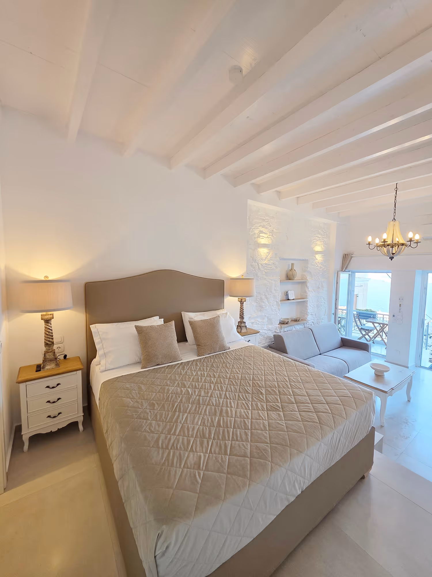Bright bedroom with beige bed, two lamps, gray sofa, white coffee table, and balcony with ocean view.