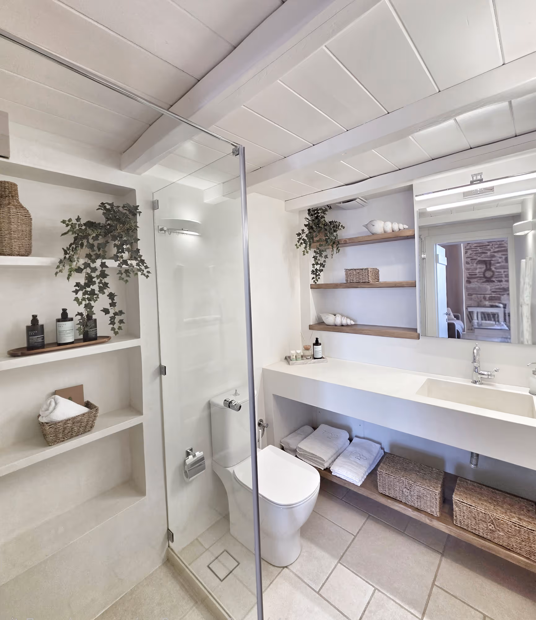 Modern white bathroom with glass shower, toilet, sink, shelves with towels, baskets, and plants.
