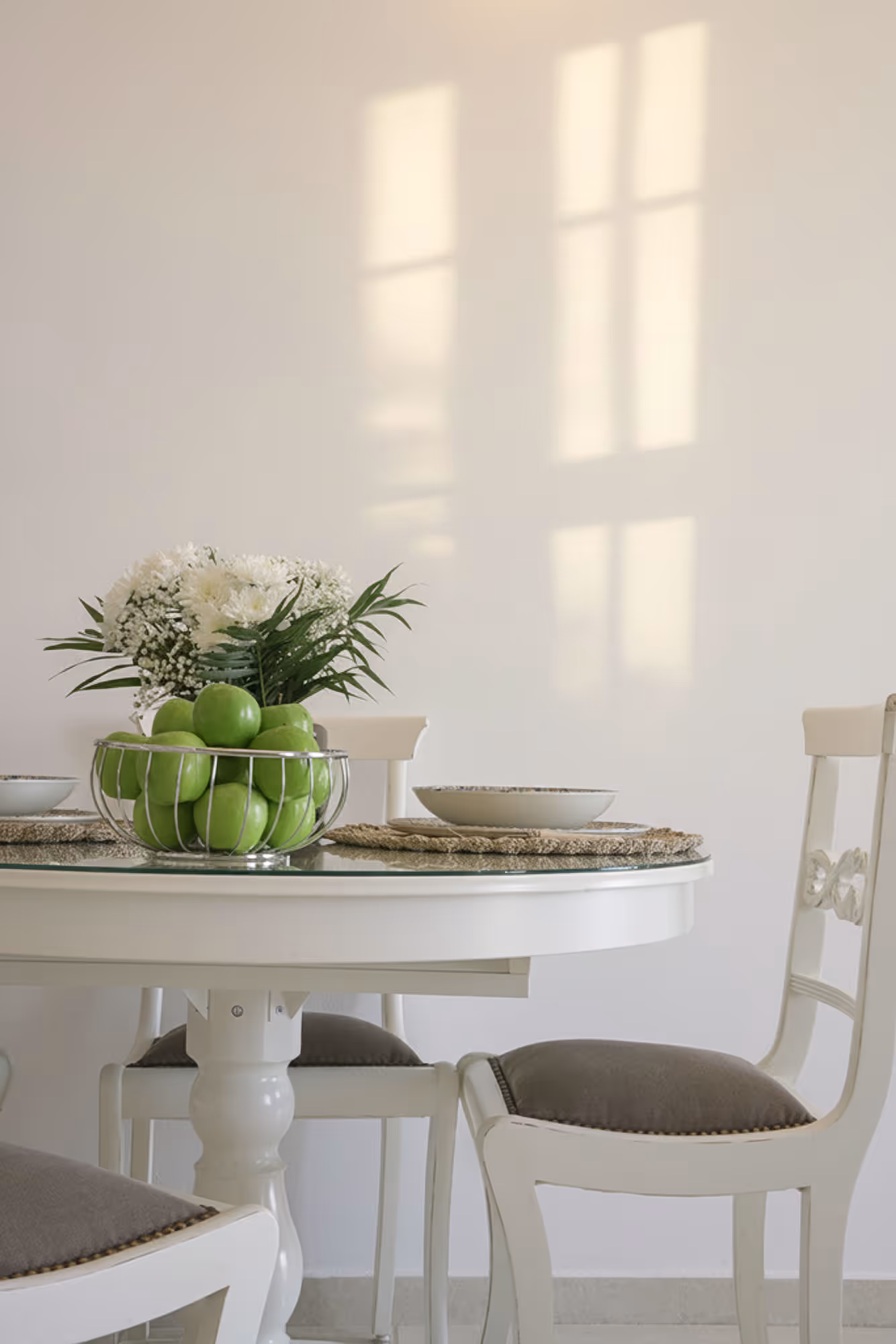White round dining table with green apples in a wire basket and white chairs with gray cushions.