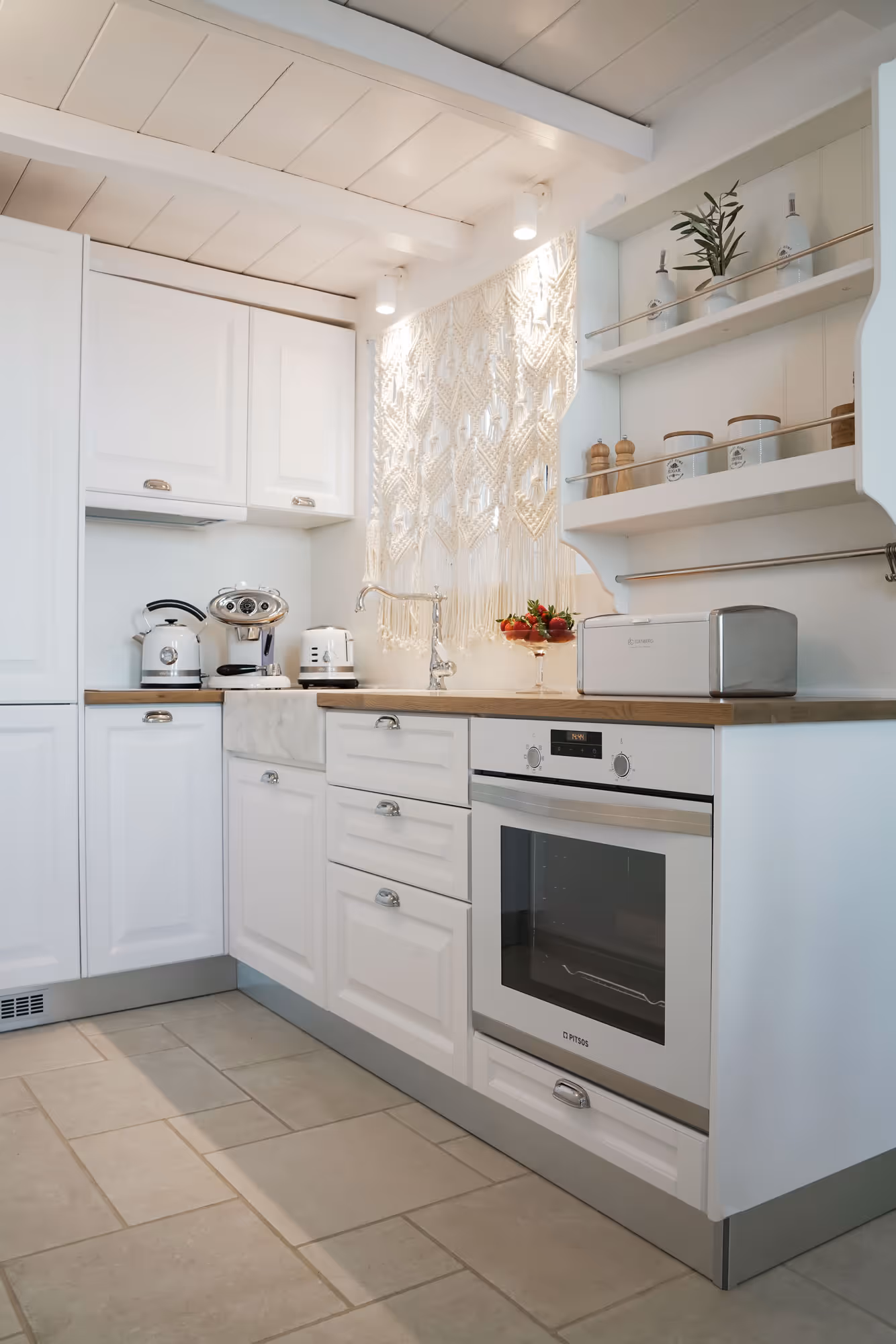 Bright modern white kitchen with wooden countertop, oven, toaster, espresso machine, and decorative macrame.