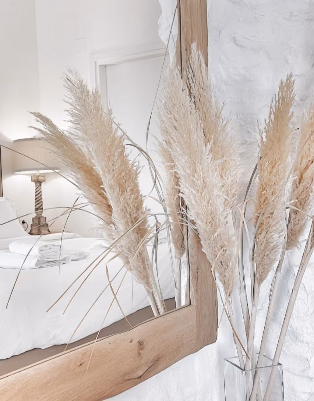 Beige pampas grass in a clear vase reflected in a large wooden-framed mirror in a white bedroom.