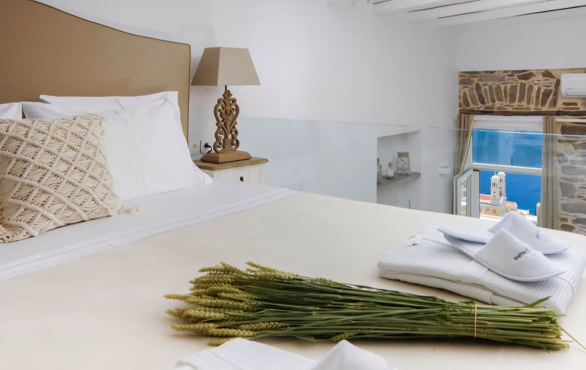 Neatly made bed with white linens, decorative pillow, bundle of green wheat, and slippers beside window with sea view.