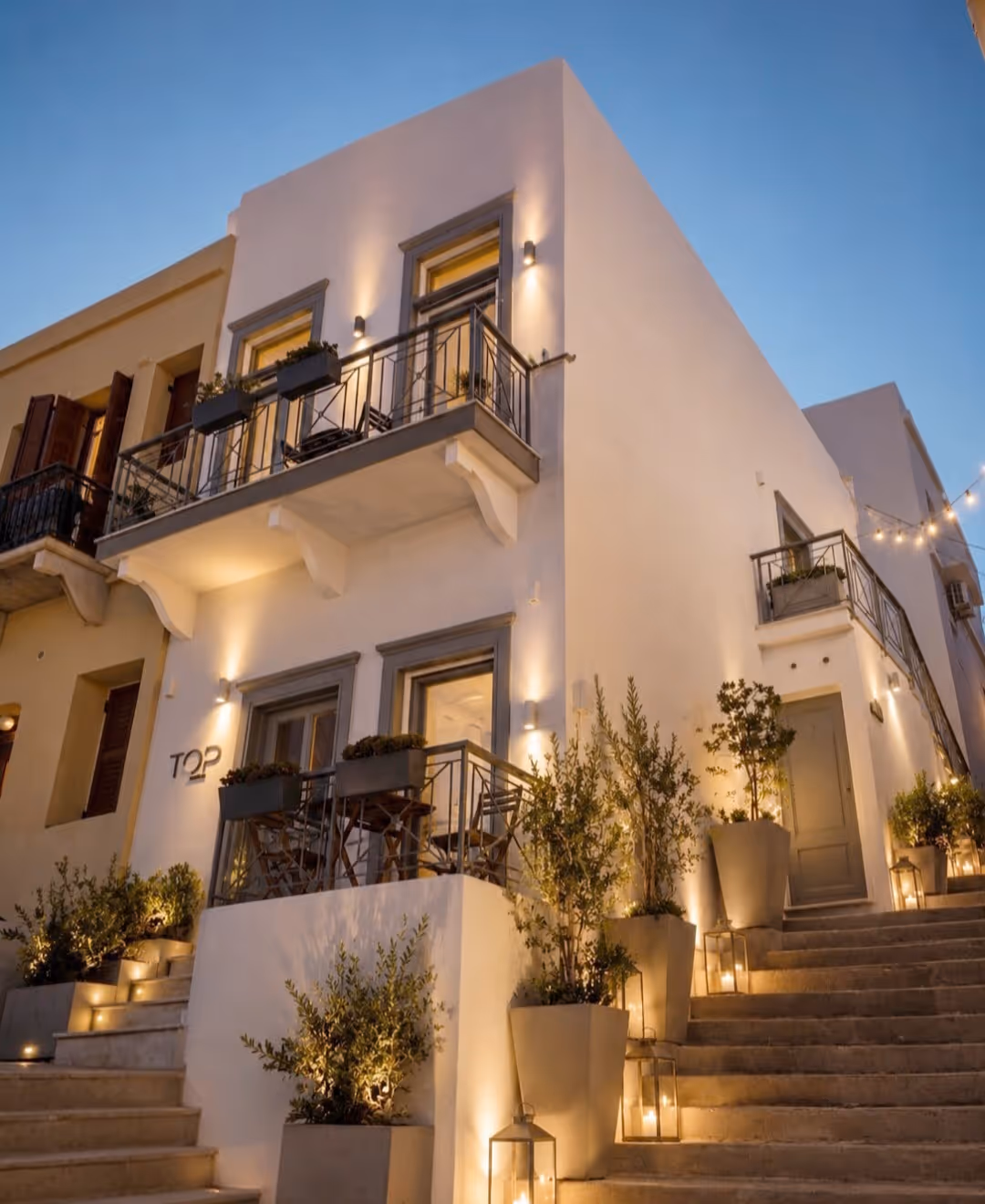 White building with balconies, plants in pots, stairs, and warm outdoor lighting at dusk.