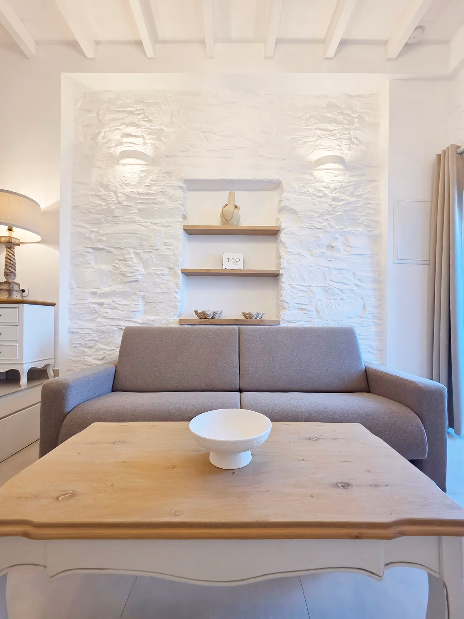 Modern living room with a gray sofa, wooden coffee table, white decorative bowl, and textured white stone wall.