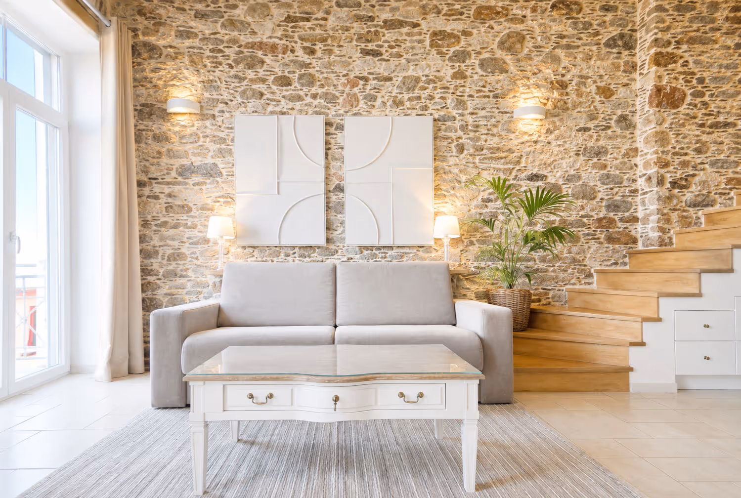 Modern living room with gray sofa, white coffee table, stone wall, wooden stairs, and indoor plant.