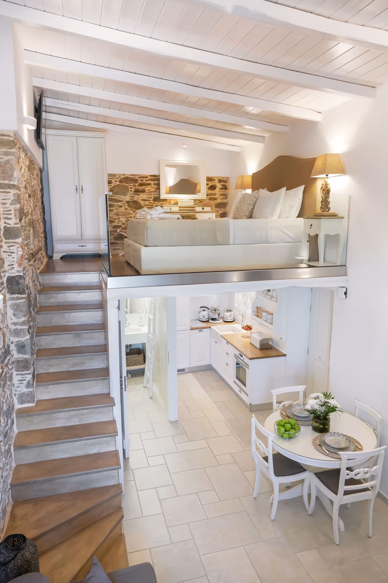 Bright loft-style apartment with elevated bed, stone wall, kitchen, dining table, and wooden stairs.