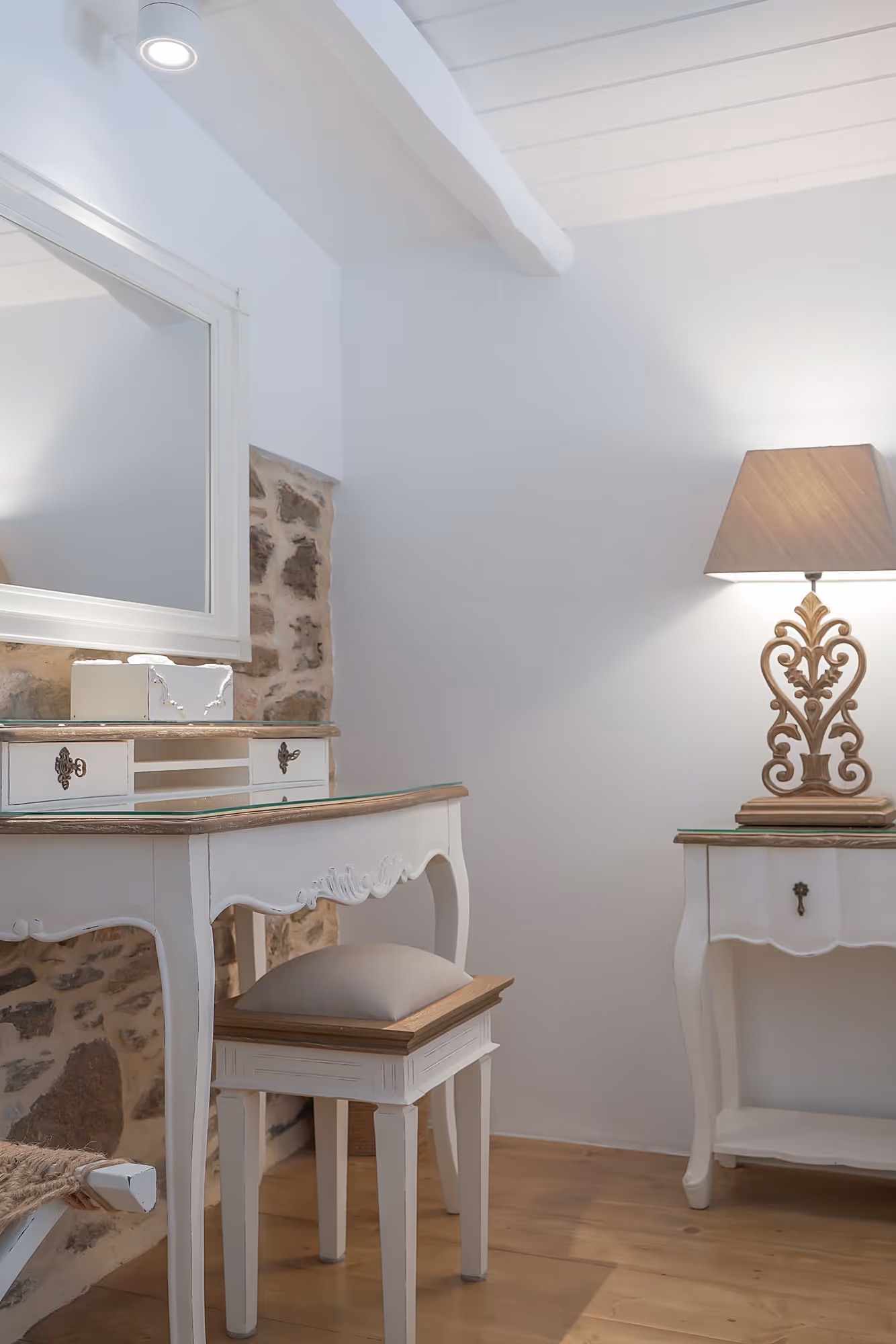 White vintage-style vanity with cushioned stool, decorative lamp, and stone accent wall in a bright room.
